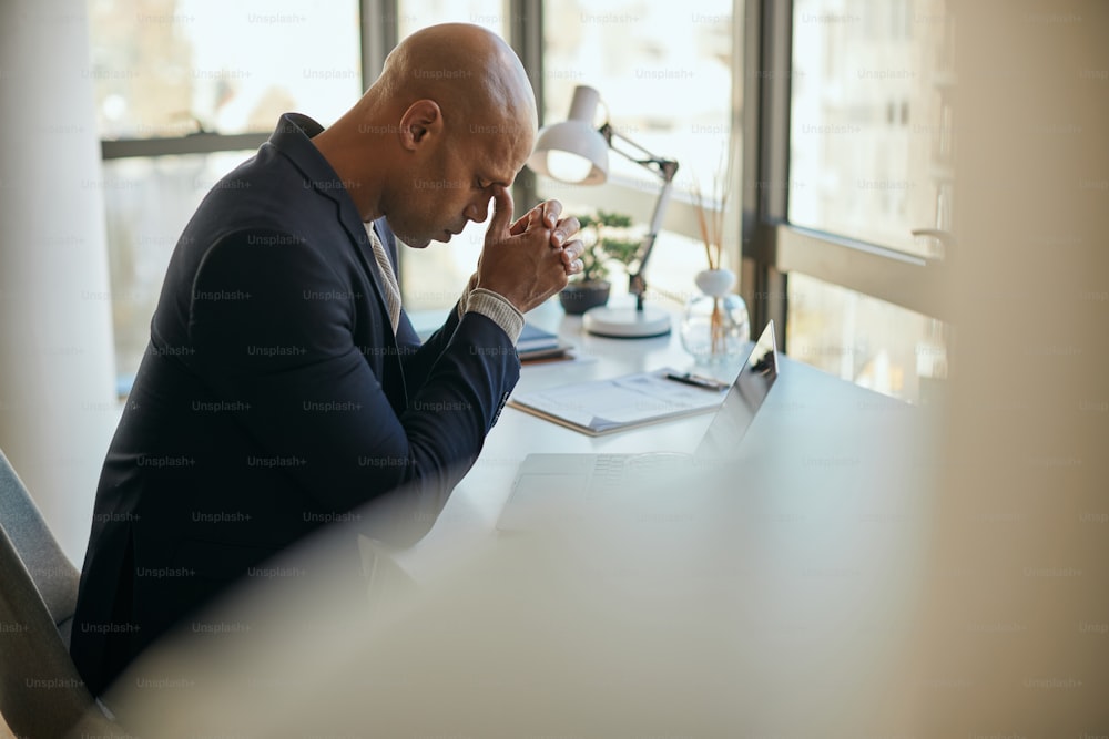 Exhausted African American businessman holding his head in pain after working on laptop in the office.