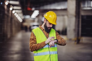 Supervisor barbudo atraente em colete, com capacete em pé dentro do estacionamento subterrâneo em processo de construção e olhando para o relógio de pulso. Os trabalhadores têm de estar a tempo para o trabalho.