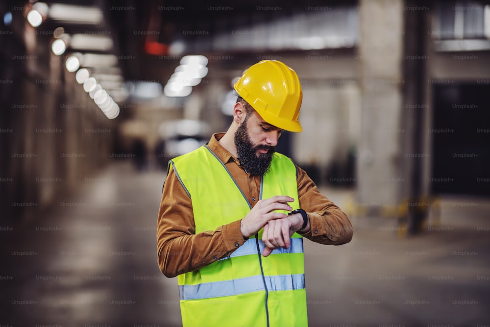 Superviseur barbu attrayant en gilet, avec un casque debout à l’intérieur d’un parking souterrain en cours de construction et regardant une montre-bracelet. Les travailleurs doivent être à l’heure au travail.