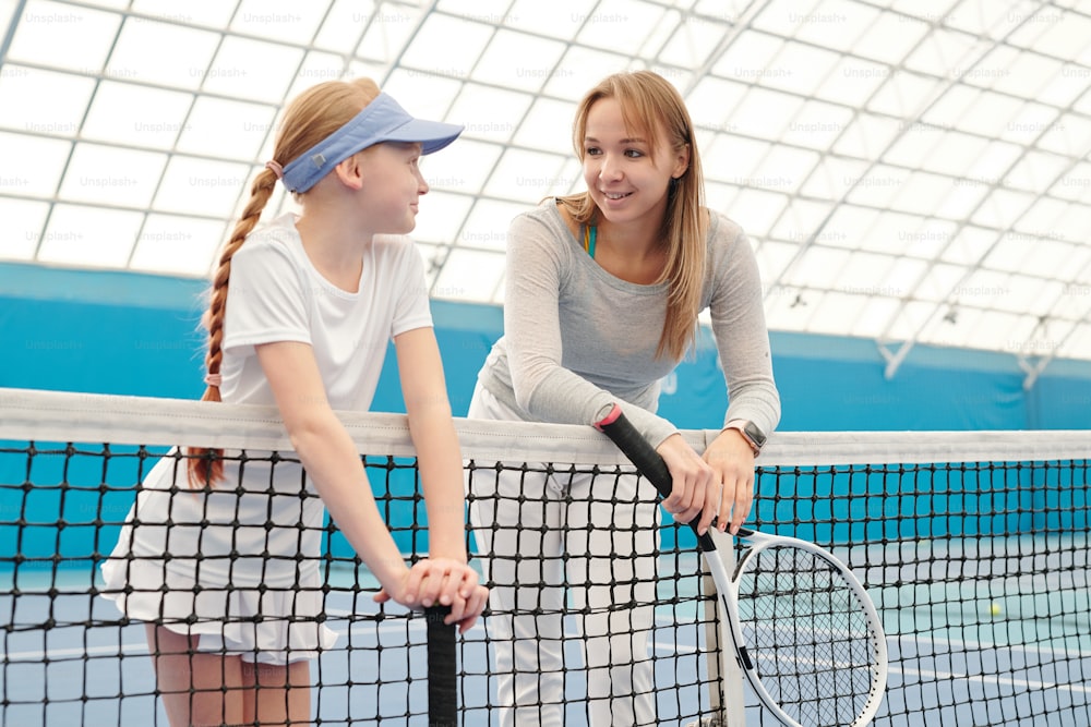 Cute blond girl in white sportswear and her trainer with tennis rackets leaning against net at stadium in front of camera and interacting