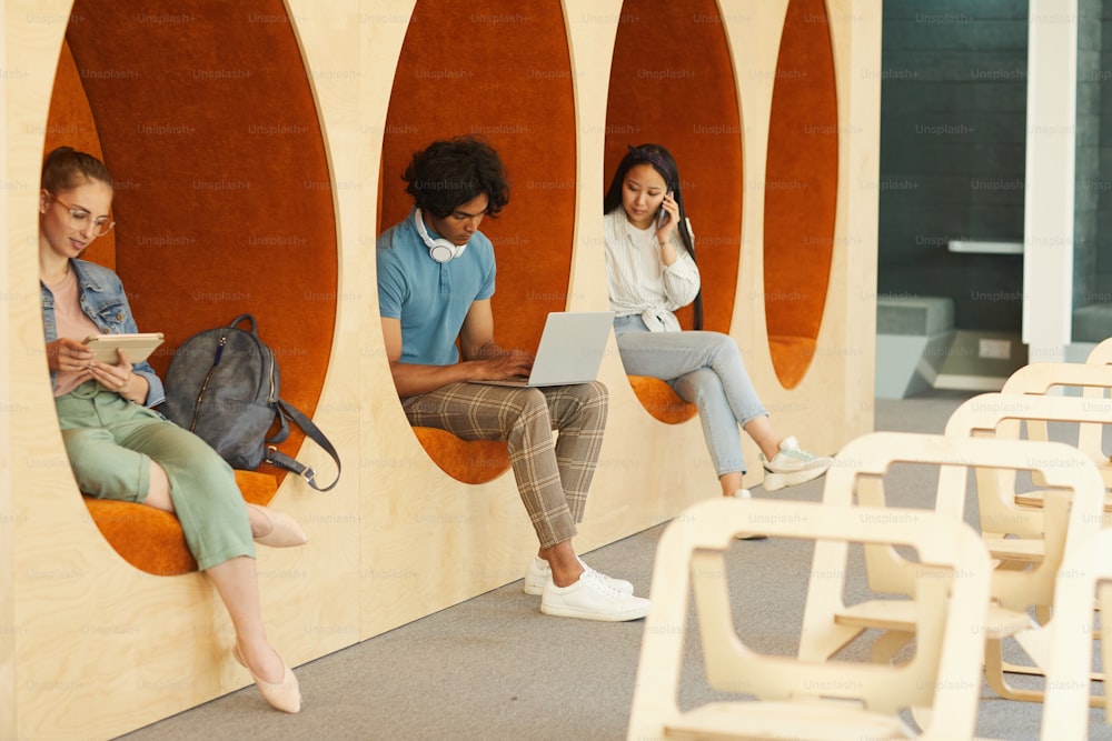 Modern multi-ethnic students sitting inside of wall chairs in university hall and using devices for education