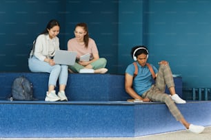 Multi-ethnic university students sitting on steps and using workbooks and devices while doing task before class in blue lobby