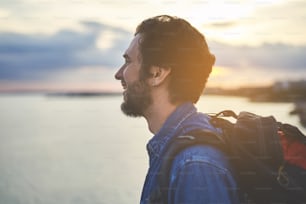 Profil eines glücklichen jungen Mannes, der die Landschaft am Meer genießt. Er steht mit Rucksack und lächelt