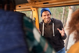 Hombre guapo parado cerca del auto y contando chistes divertidos a sus amigos. El bosque borroso al fondo