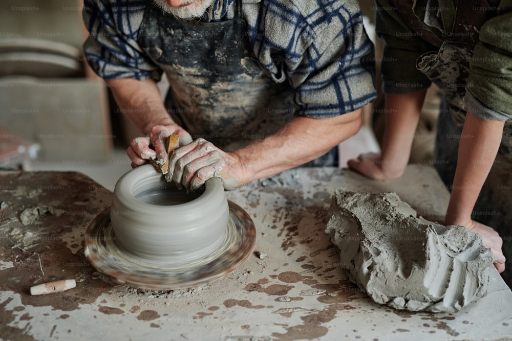 Primer plano del artista de cerámica que da forma a la arcilla en la rueda de alfarería en el taller con su asistente