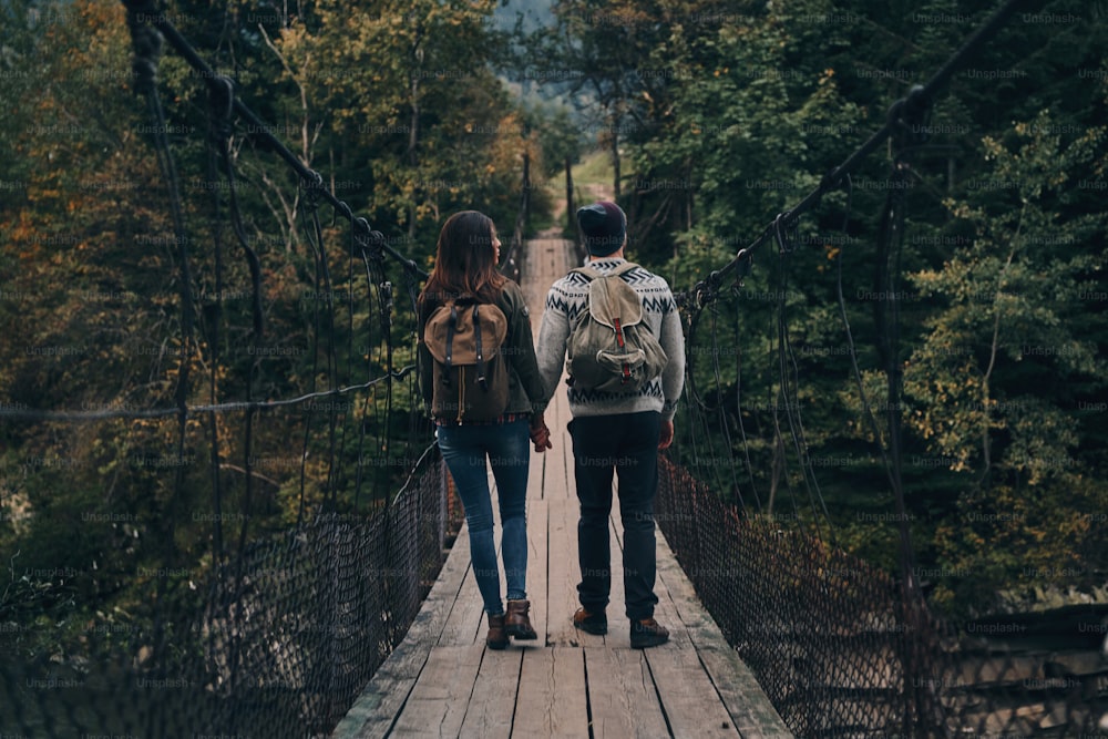 Vista trasera de cuerpo entero de una pareja joven tomada de la mano mientras camina por el puente colgante