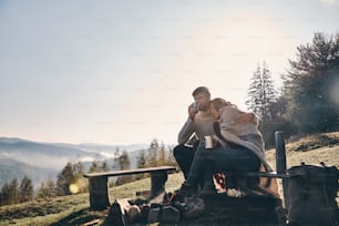 산에서 모닥불 옆에 앉아 모닝 커피를 마시는 아름다운 젊은 커플