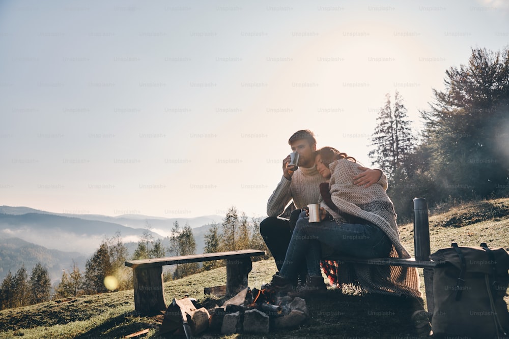 Beautiful young couple having morning coffee while sitting by the campfire in mountains