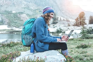 Young beautiful traveling girl with backpack, hat and laptop sitting on rock and trying to connect network in the stunning mountain wilderness in front of amazing cold lake.