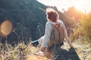 Young woman with backpack sitting on a cliff and enjoying the view of mountains, sunset and landscape.