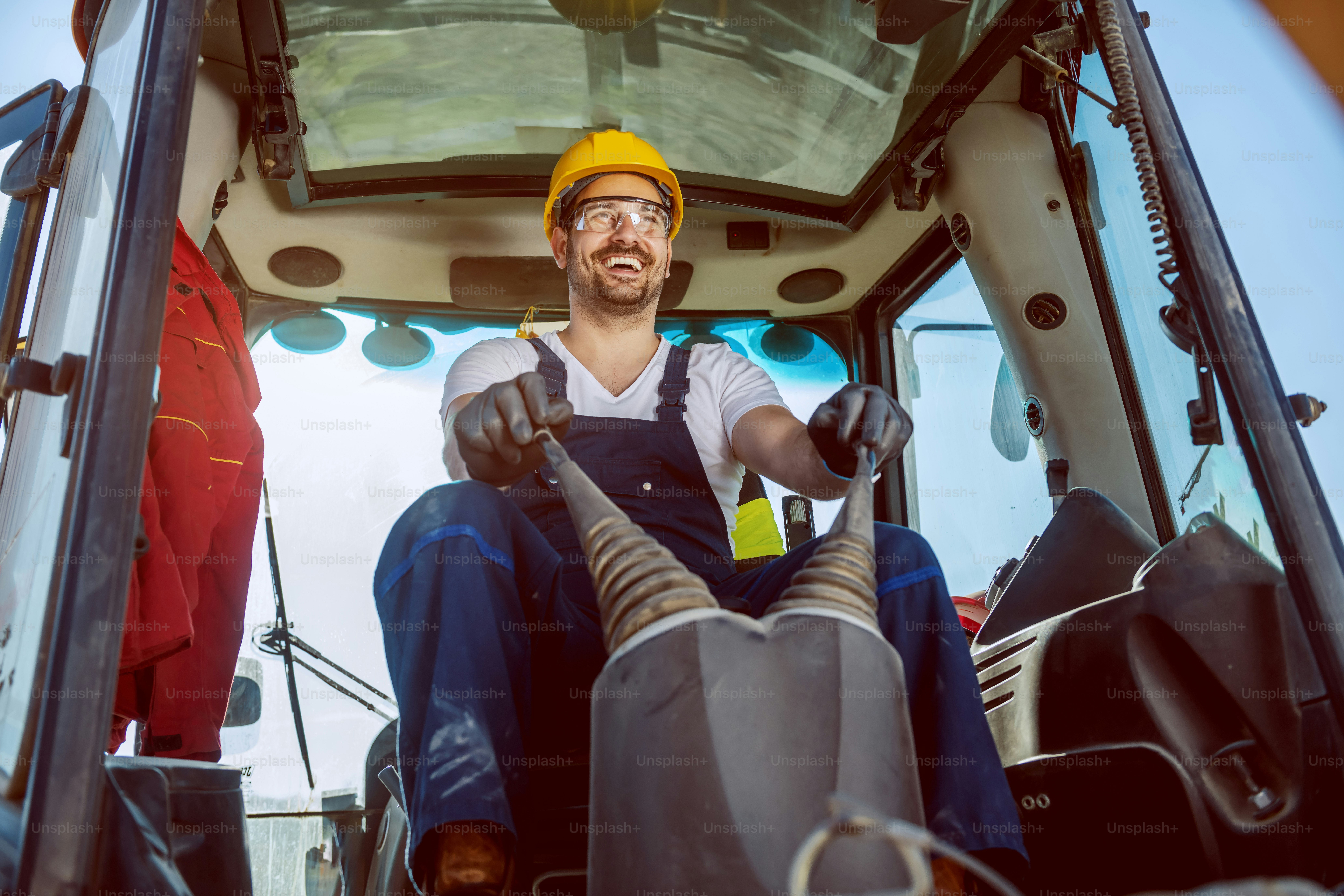 Sorridente bel lavoratore caucasico in tuta e con casco in testa che guida l'escavatore.