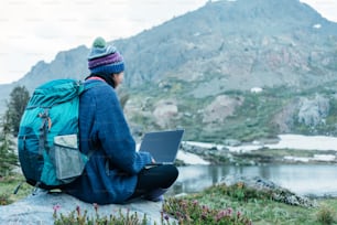 Young traveling woman with backpack and hat sitting on rock and using laptop to connect network in the stunning mountain wilderness in front of amazing cold lake.