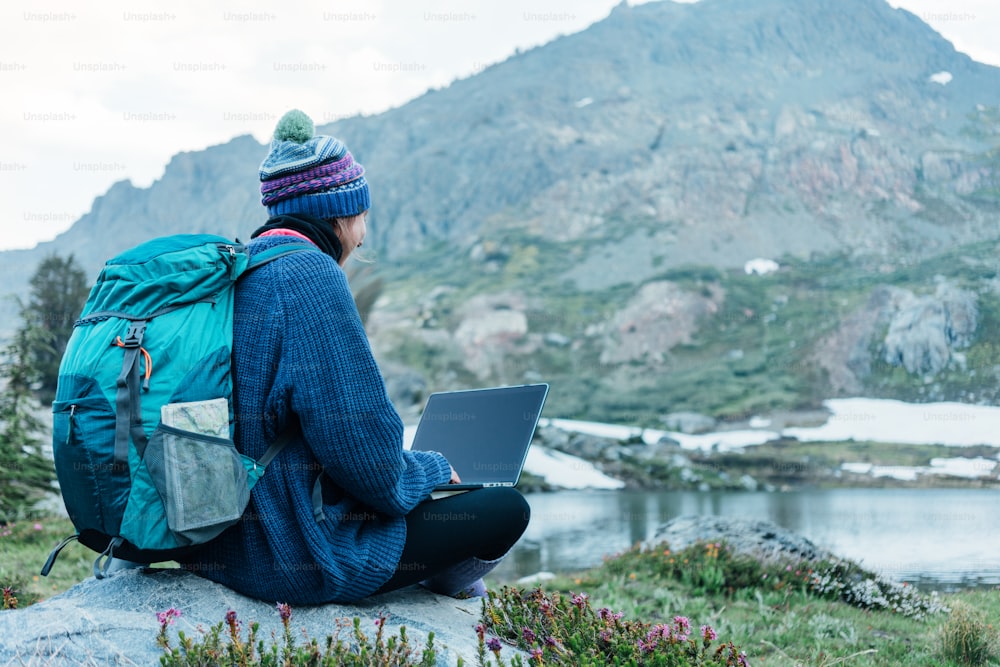 Young traveling woman with backpack and hat sitting on rock and using laptop to connect network in the stunning mountain wilderness in front of amazing cold lake.