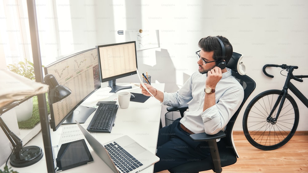 Talking about business. Side view of young bearded trader in headset holding financial report and talking with client while sitting in front of monitor screens in the office. Business concept. Trade concept. Call center