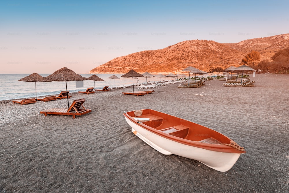 Sunbeds and sun umbrellas await vacationers on the shingle beach at Ovabuku beach on the Datca Peninsula in Turkey. Tourist boat at the foreground