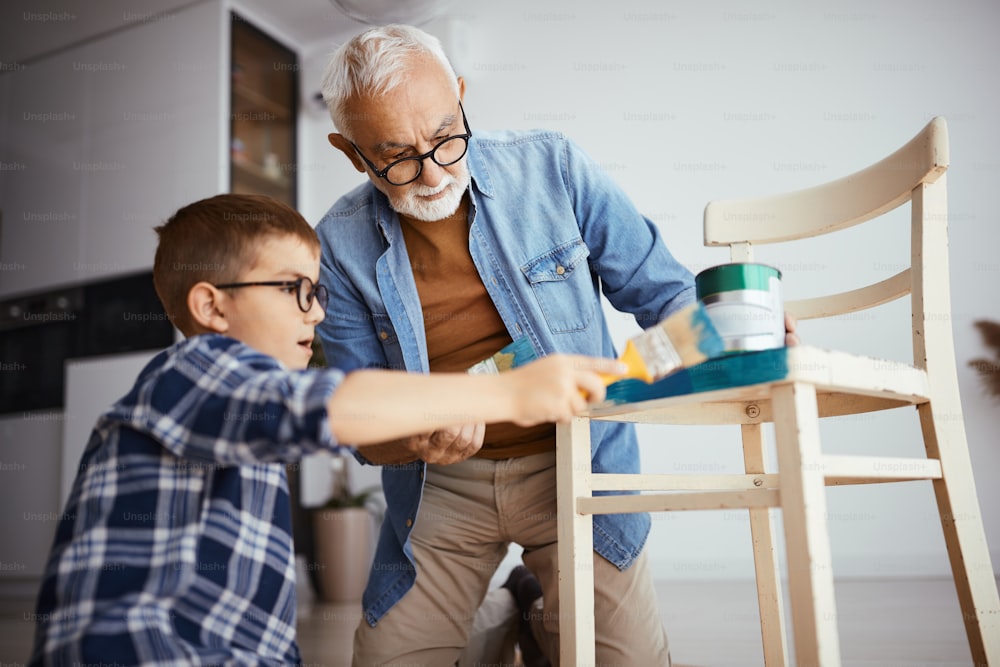 Senior man and his grandson painting wooden chair in blue color at home.
