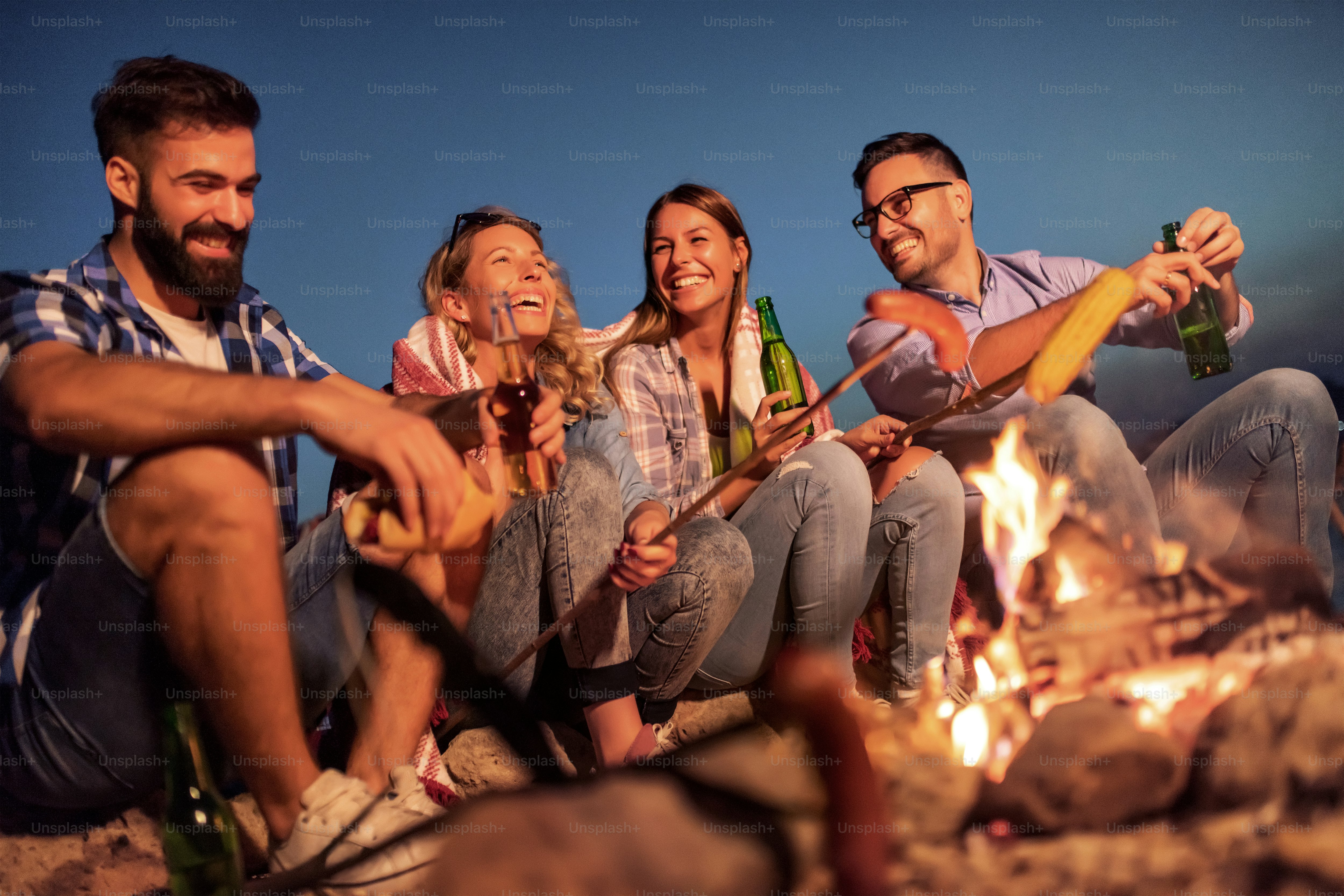 Grupo de amigos sentados à beira do fogo à noite, grelhando salsichas e se divertindo muito na praia.