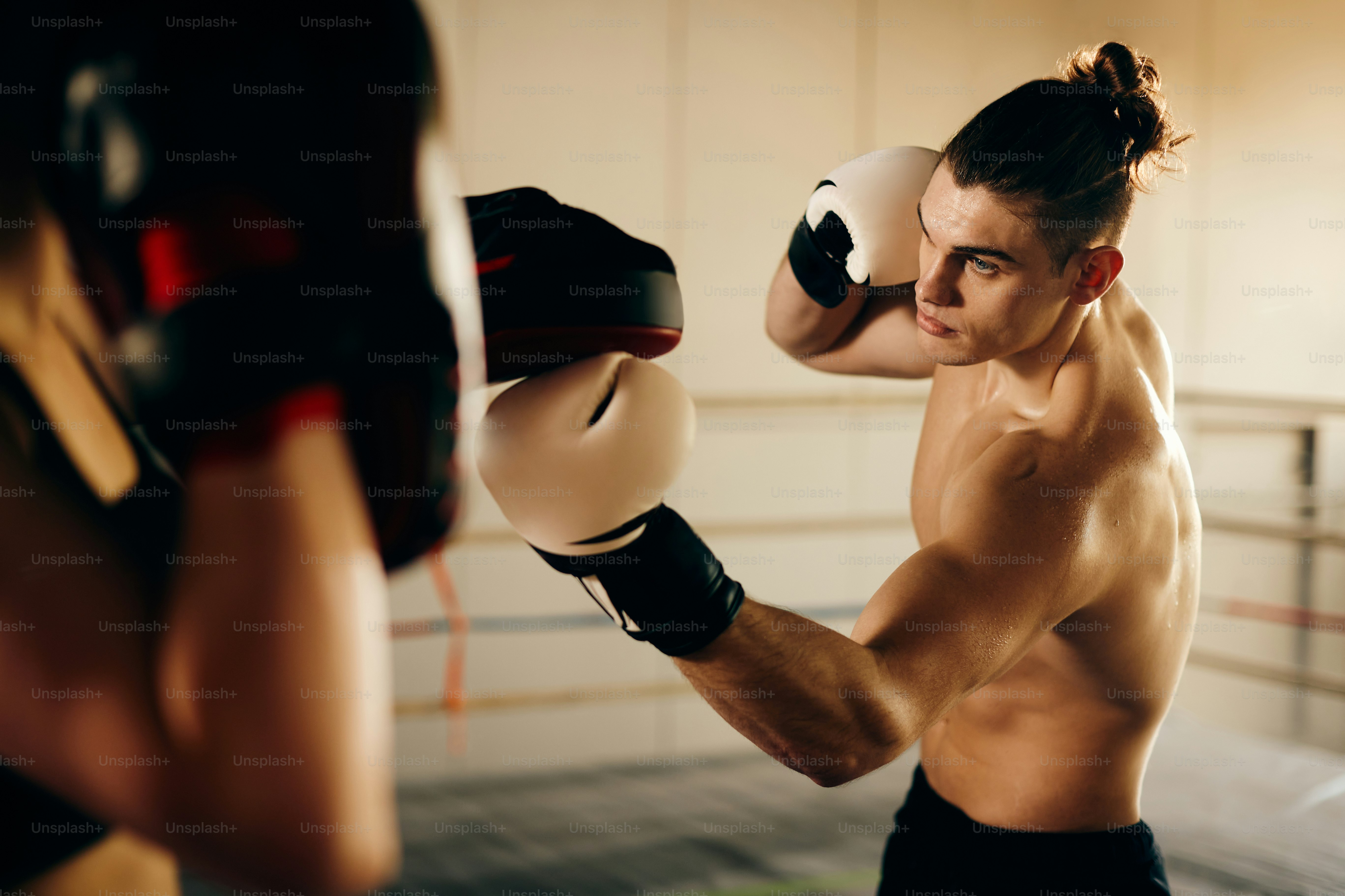 Shirtless fighter practicing boxing with his coach in a gym.