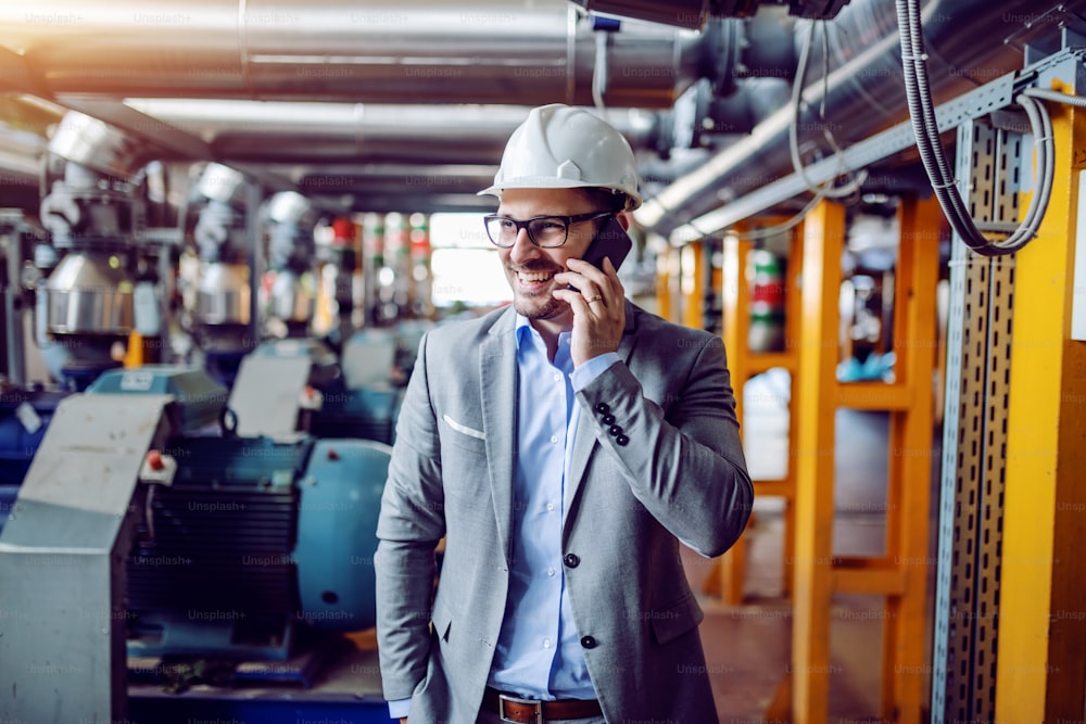 Attractive smiling caucasian businessman in suit and with helmet on head talking on the phone while standing in power plant.