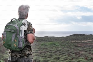 Back view of senior adult man with backpack on shoulders enjoying outdoors excursion between green bushes and sea. A white-haired elderly people in healthy activity under the rain