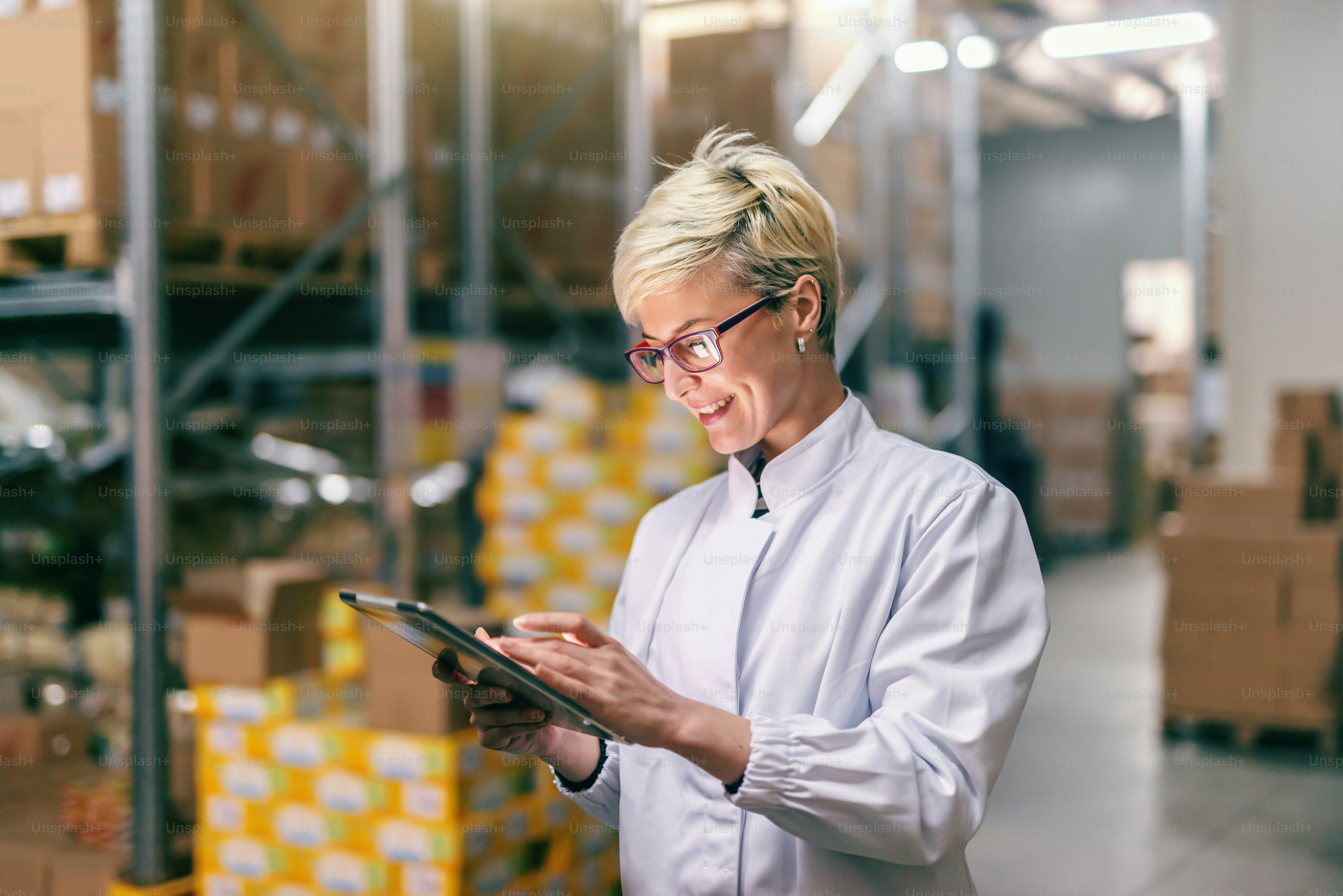 Young Caucasian blonde woman in white uniform using tablet in warehouse.