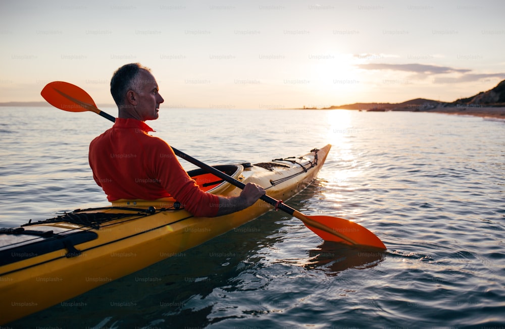 Vista posteriore dell'uomo anziano attivo che si diverte a pagaiare in kayak sul mare al tramonto.