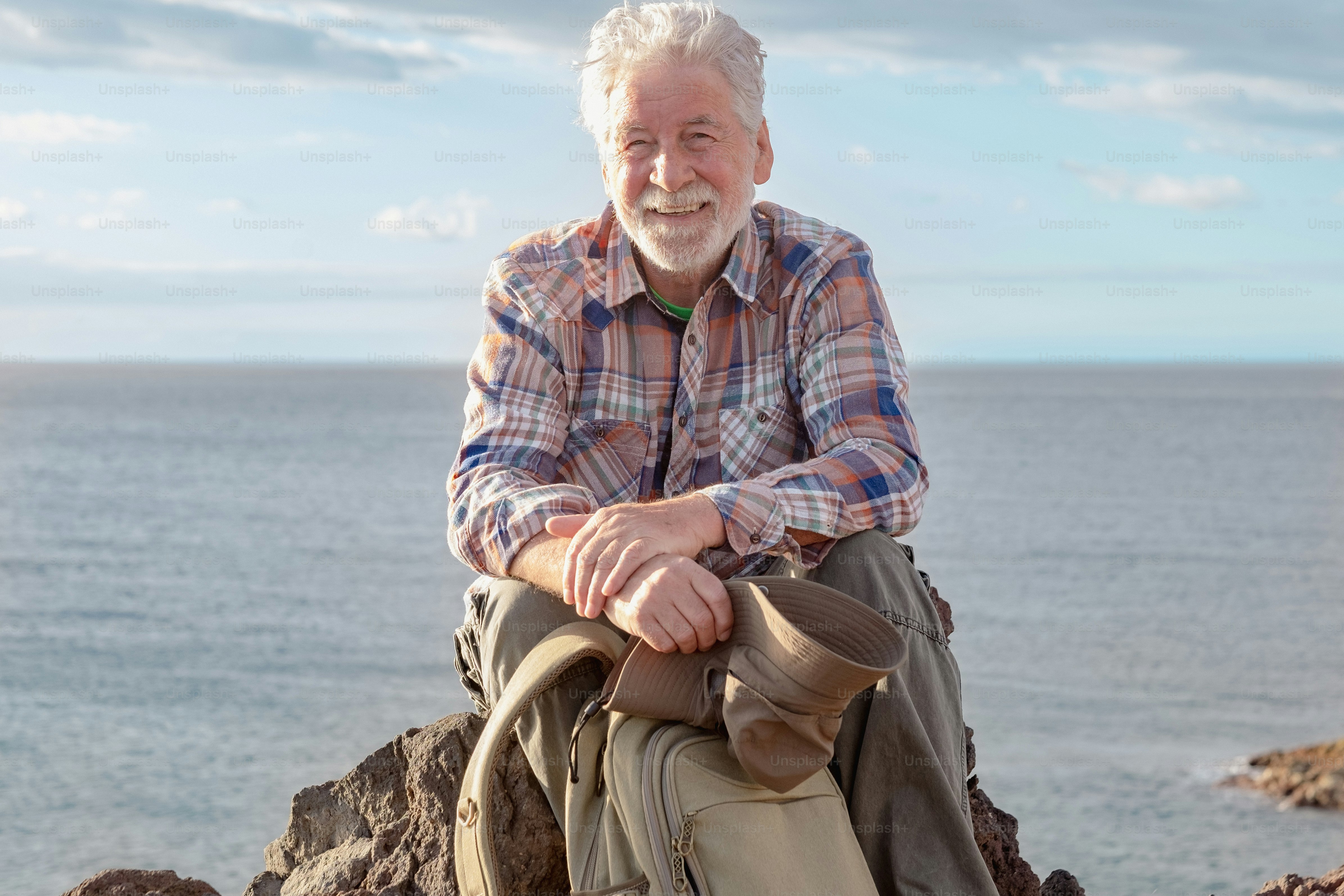 Portrait of senior smiling man resting in outdoor excursion at sea sitting on the cliff looking at camera. Mature attractive bearded man on a hiking trip enjoying freedom and healthy vacation