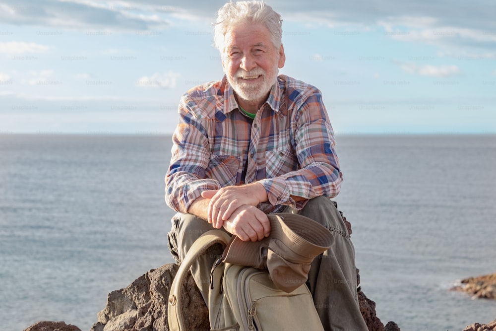 Portrait of senior smiling man resting in outdoor excursion at sea sitting on the cliff looking at camera. Mature attractive bearded man on a hiking trip enjoying freedom and healthy vacation