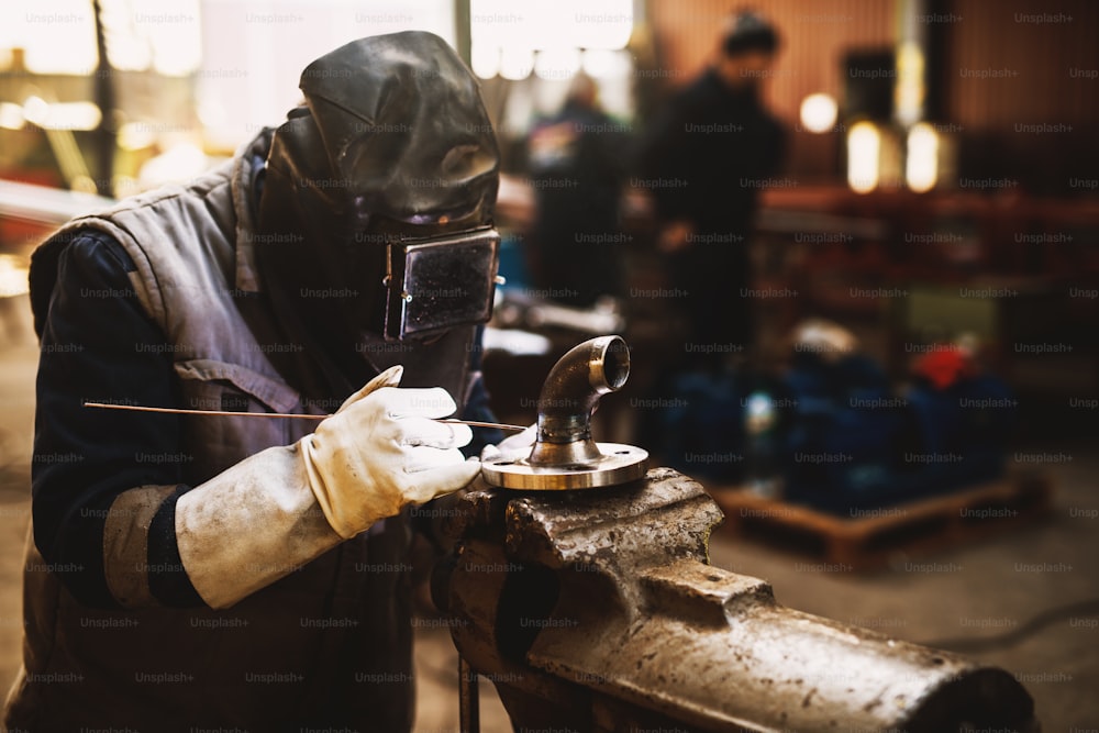 Welder in protective uniform and mask welding metal pipe on the industrial table while sparks flying.