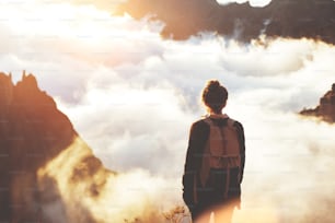 Young traveler girl with backpack standing on edge of cliff and enjoying view of mountains, sunset and clouds.