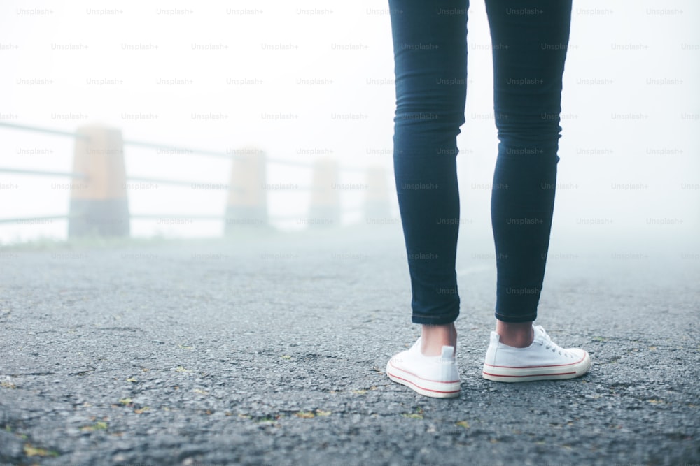 Travelling woman wearing jeans and sneakers standing on the road in fog close-up.