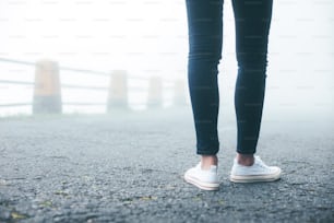 Travelling woman wearing jeans and sneakers standing on the road in fog close-up.