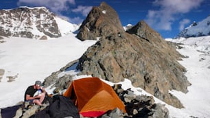 남자 산악인 등반가가 생모리츠 근처 스위스 알프스에서 주변의 멋진 산 풍경이 있는 주황색 텐트 옆에서 베이스캠프에서 식사를 하고 있다.
