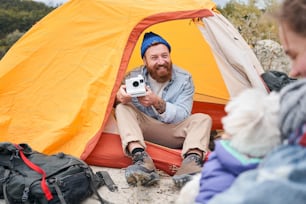 Bel homme barbu souriant et prenant des photos sur le polaroid de sa femme bien-aimée avec un chien dans ses bras alors qu’elle est assise dans une tente