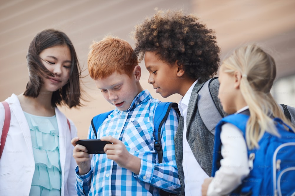 Gruppe von Freunden, die stehen und auf das Handy ihres Klassenkameraden schauen, während er darin spielt, sie stehen nach dem Schulunterricht im Freien