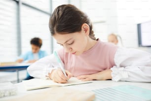 Caucasian female middle schooler with dark brown hair sitting at school desk writing essay, horizontal medium close up portrait