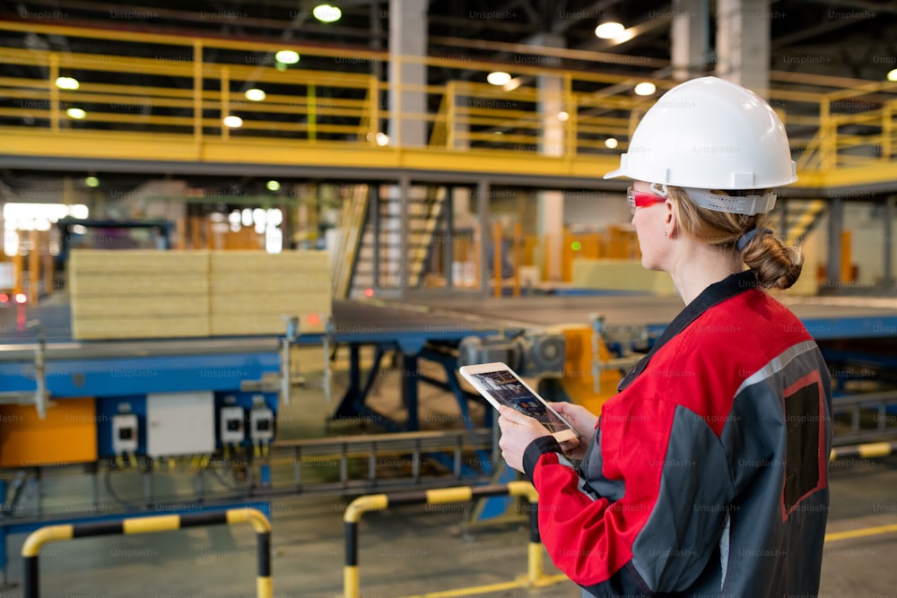 Vue arrière d’une femme occupée portant un casque de sécurité et des lunettes de sécurité à l’aide d’une tablette tout en contrôlant le processus de la chaîne de production à l’usine