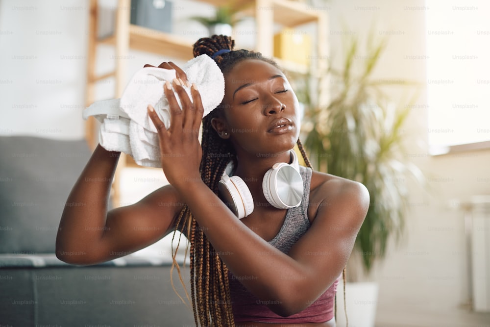 Deportista negra exhausta usando una toalla mientras se limpia el sudor de la cara después de hacer ejercicio en casa.