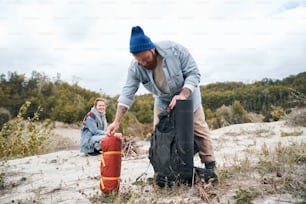 Bearded hipster man pulling a tent out of a large tourist backpack to preparing for a halt while his girlfriend making a campfire