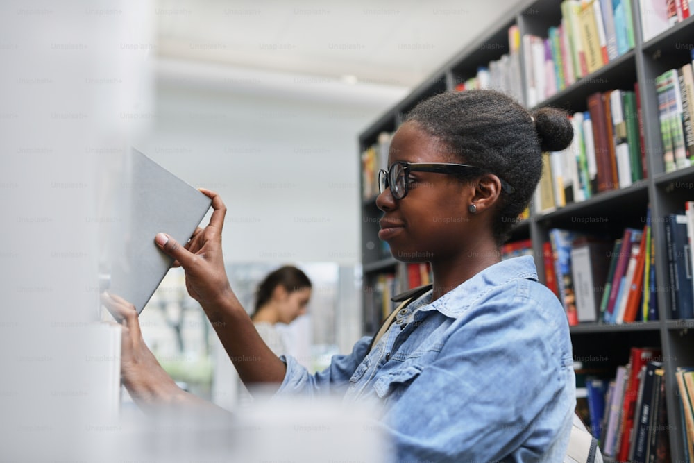 African young woman in eyeglasses taking book from the bookshelf while standing in the library