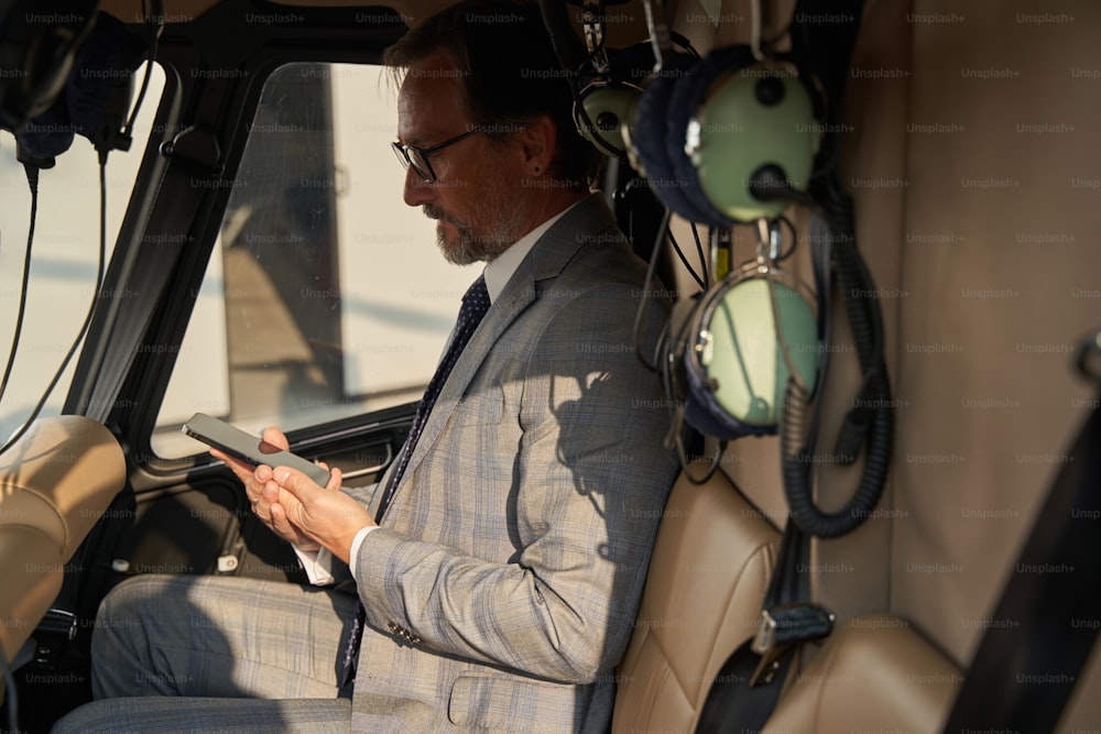 Side view of gentleman seated in helicopter cockpit staring at cellphone in hands