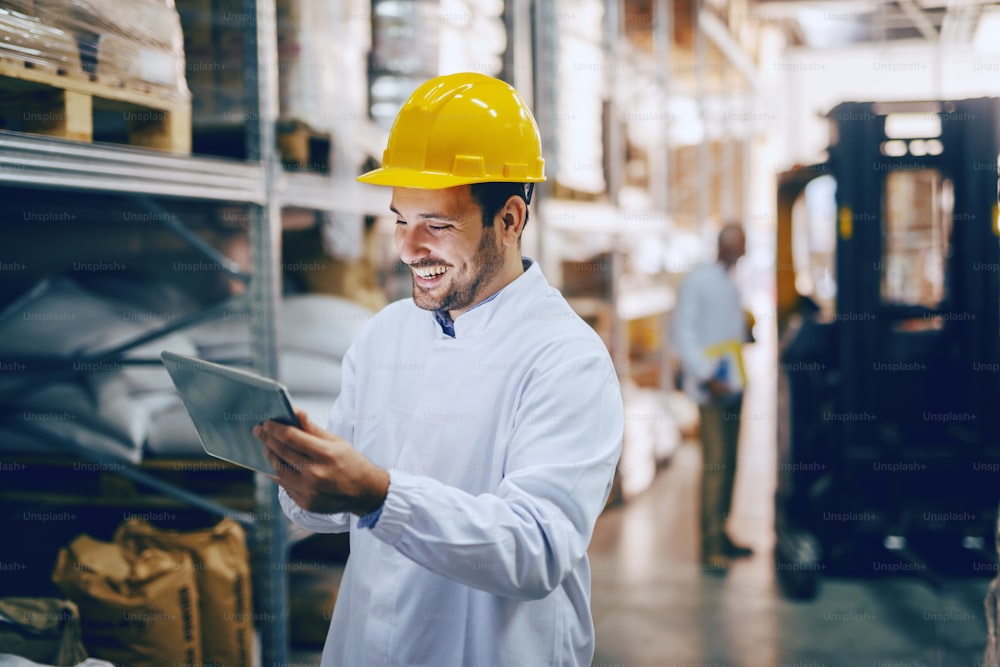 Young smiling Caucasian auditor in white uniform and with yellow helmet on head using tablet for checking on goods in warehouse. In background worker standing next to forklift.