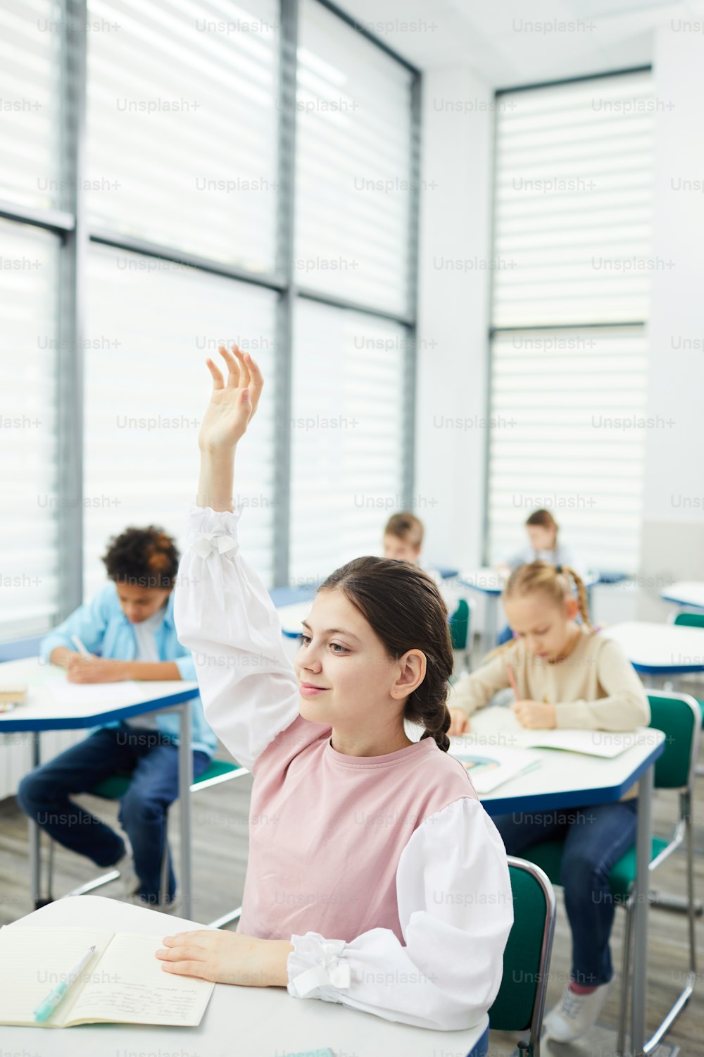 Vertical portrait of active smart twelve-year-old girl with dark hair sitting at school desk raising her hand in class, copy space