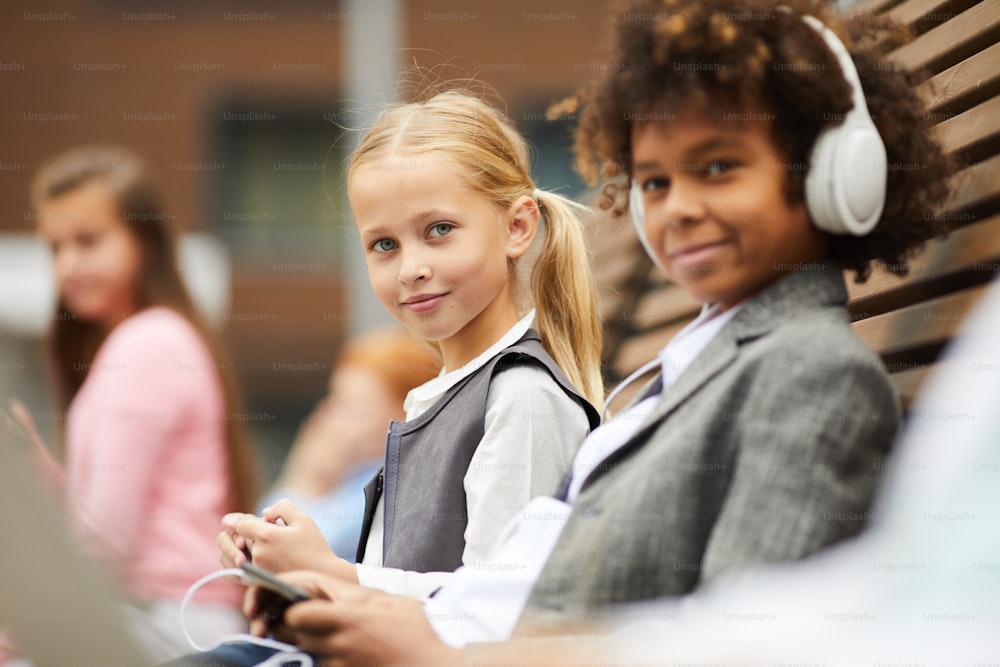 Porträt einer süßen Schülerin mit blonden Haaren, die in die Kamera schaut, während sie zusammen mit ihrer Klassenkameradin, die ein Mobiltelefon benutzt, auf der Bank sitzt