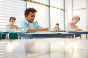 Horizontal shot of young middle schoolers wearing casual clothes sitting at desks in modern classroom doing lesson exercises, copy space