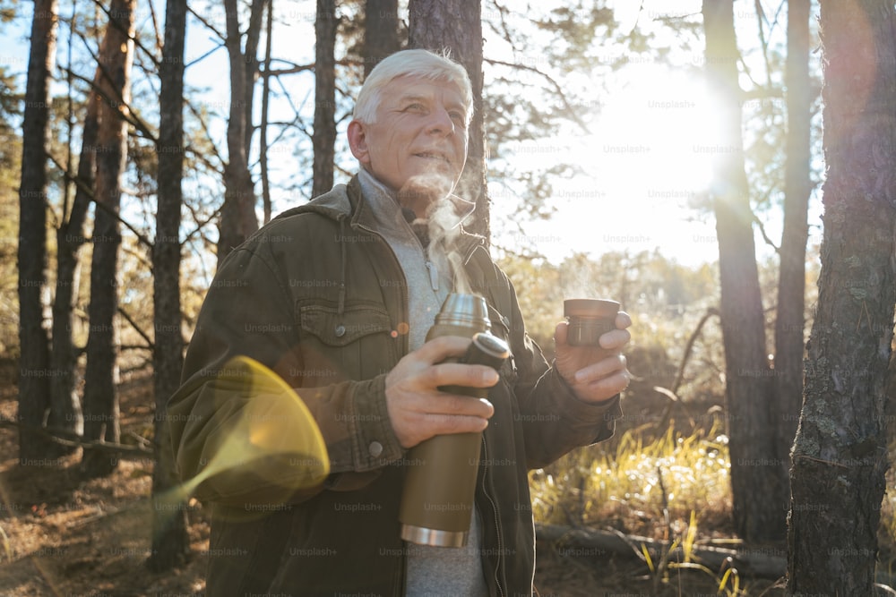 차가운 가을 숲을 산책 한 후 휴식을 취하면서 먼 곳을 바라보고 뜨거운 차를 마시는 성숙한 남자의 허리 위로 초상화. 스톡 사진