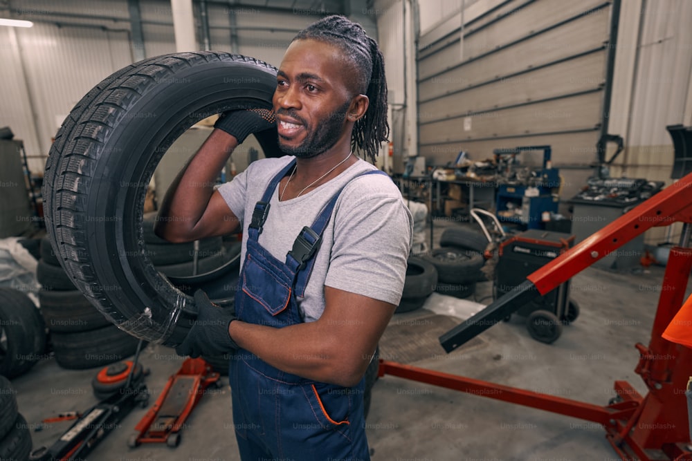 Mecánico masculino en taller de reparación que lleva el neumático al coche con la mano derecha