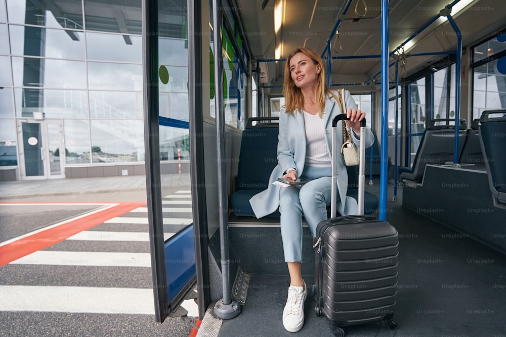 Calm fashionable lady with trolley suitcase sitting in parked airport shuttle bus