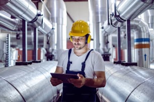 Focused plant worker in overalls, with protective helmet on head and antiphons on ears using tablet for checking machine.