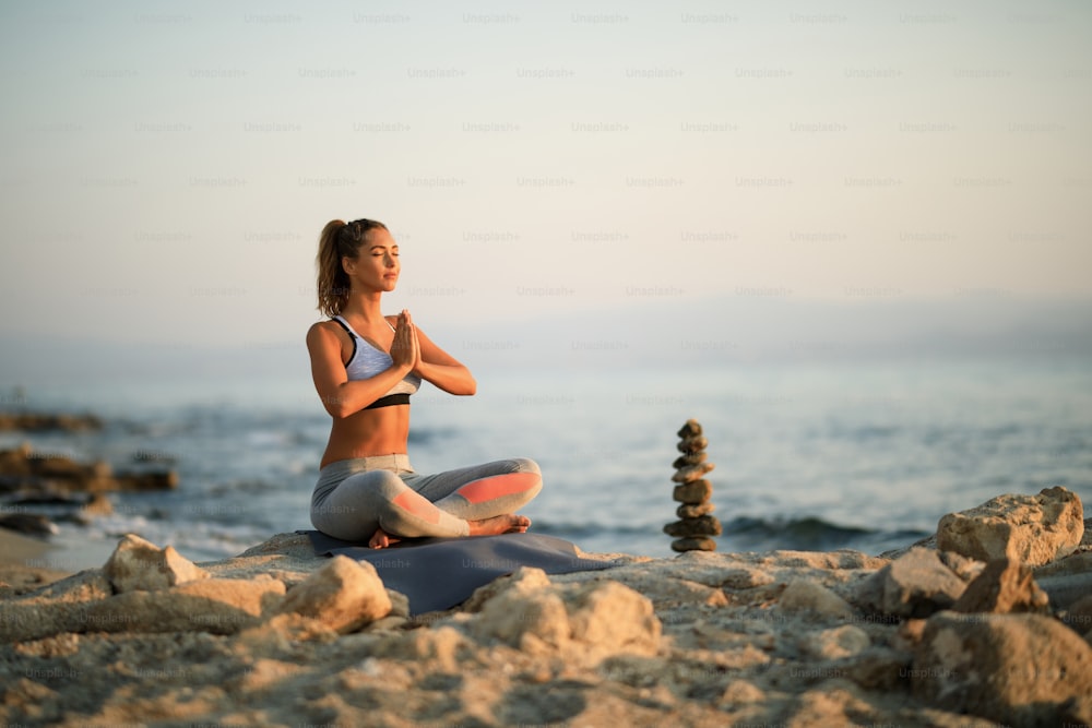 Young woman with hands clasped meditating on beach rock during morning Yoga class. Copy space.